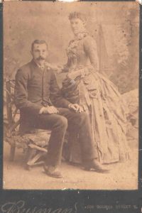 Historic portrait photo of husband and wife wearing suit and elaborate dress. Man is seated woman is standing.