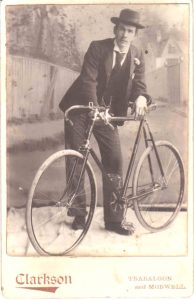 Historic photo of a man wearing a suit with a hat, leaning against a bicycle