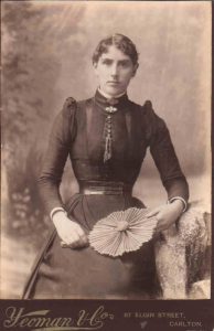 Historic portrait photo of a young woman in dark dress with a tight corset (she has a small waist) holding a fan