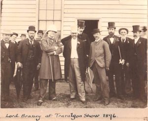 Historic photo of Lord Brassey and 11 men in suits and top hats, standing infront of wooden building, at the Traralgon Show in 1896