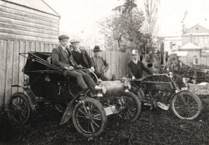 Men sitting in Oldesmobile and standing near a motorised bike in 1903