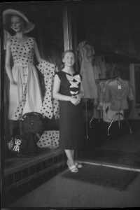 Black and white photo of woman in black dress with large polka-dot bows down the front, standing at entry to a fashion boutique in 1957