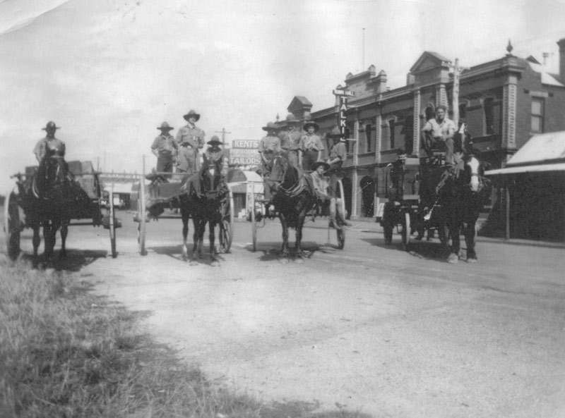 A scanned copy of a sepia photographic print of four horse-drewn carts, carrying Boy Scouts, in Hotham Street outside the Traralgon Town Hall.